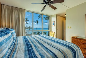 A bedroom with a large bed, a ceiling fan, a window with a view of palm trees and the ocean, and a wooden dresser.