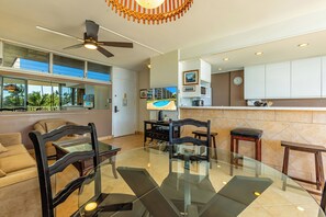 A modern living room with a glass dining table, black chairs, a ceiling fan, and a view of the pool through large windows.
