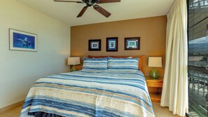 A bedroom with a large bed, a ceiling fan, framed pictures on the wall, and a window with curtains. - Island Paradise Found: Comfort and Oceanfront Views at Kamaole Nalu #404 (Kihei)