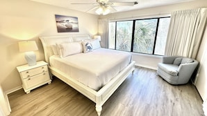 A bedroom with a large bed, a nightstand, a chair, a ceiling fan, and a window with curtains.