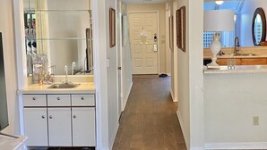 A hallway with a bathroom on the left, featuring a glass-enclosed vanity and a sink. A door leads to a bedroom with a bed and a nightstand. The hallway has wooden flooring and white walls.