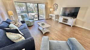 A living room with a large TV, a sofa set, a coffee table, and a view through the glass doors.