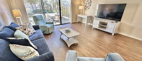 Uma sala de estar com uma TV grande, um conjunto de sofá, uma mesa de centro e vista através das portas de vidro.