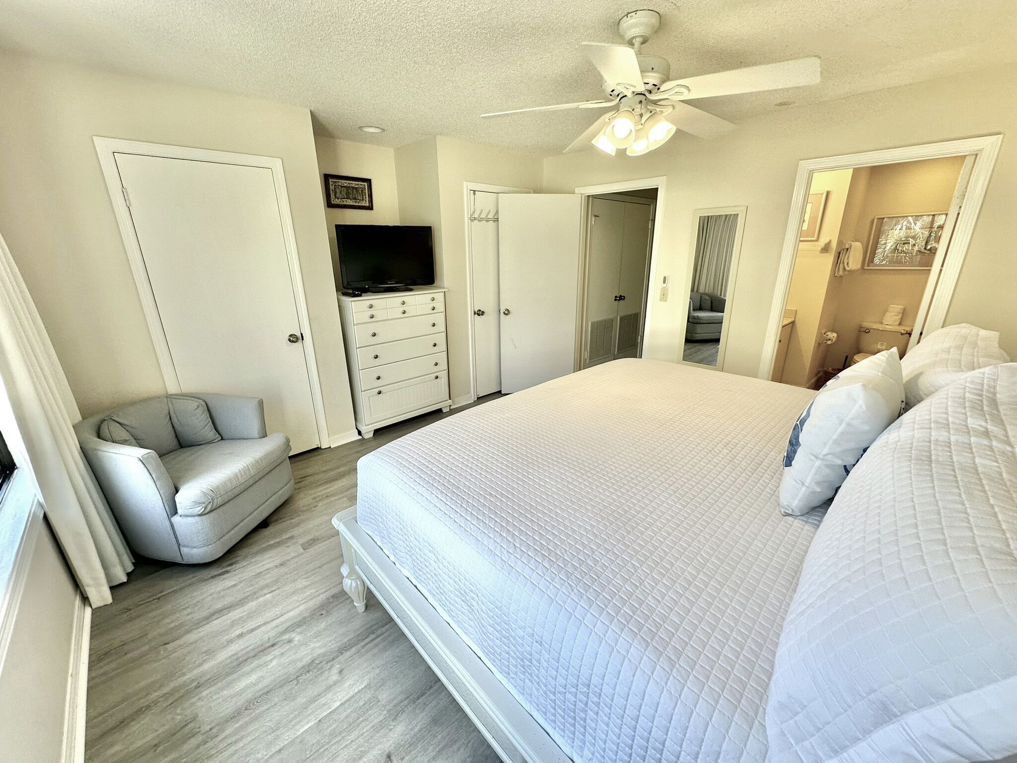 A bedroom with a large bed, a ceiling fan, a TV on a dresser, and a gray armchair.