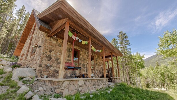 A stone building with a wooden porch, surrounded by trees and a mountain in the background. - Monarch Crest Chalet- Sauna and Pet Friendly (Salida)