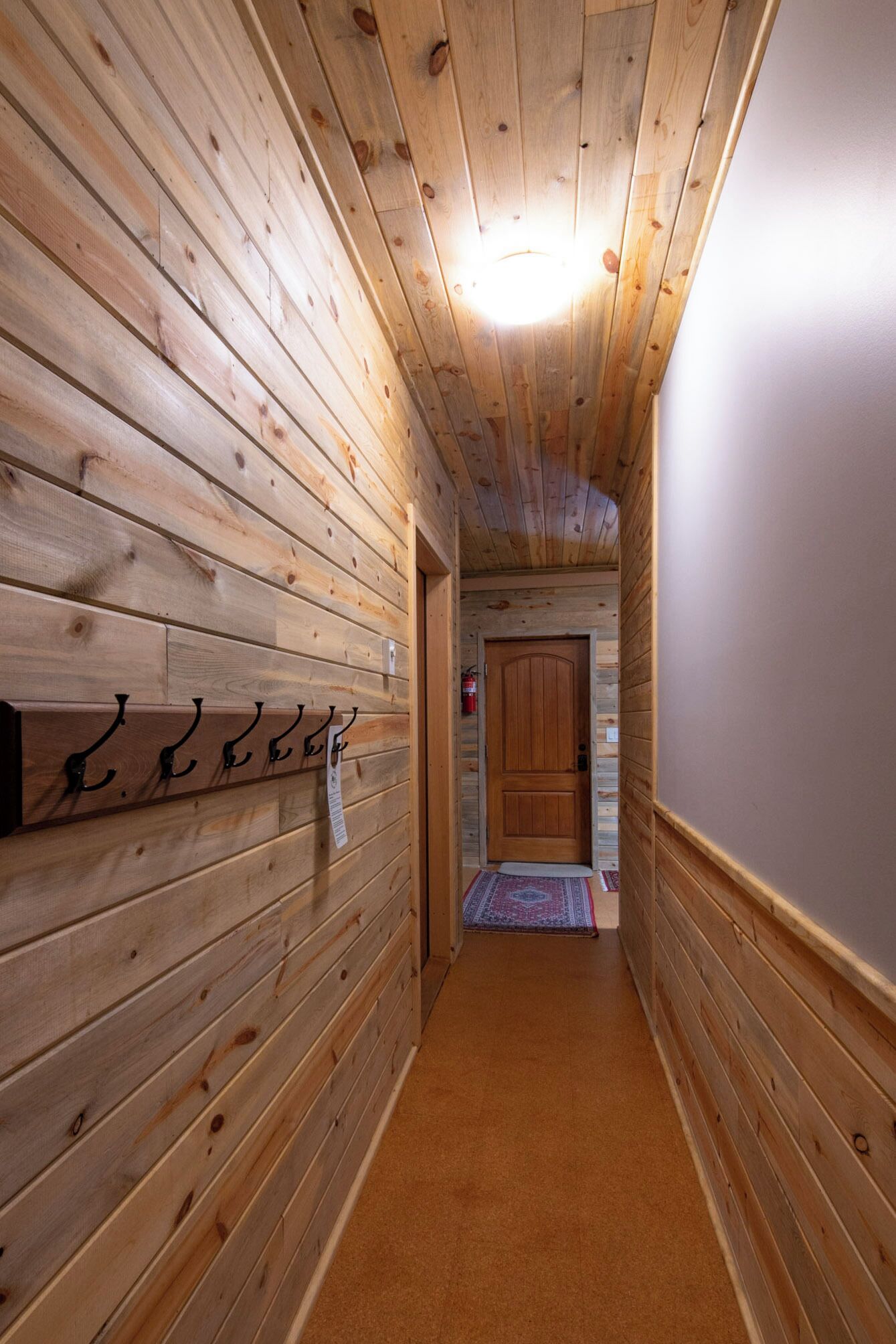 A wooden hallway with a wooden door at the end, a coat rack with hooks, and a fire extinguisher on the wall.