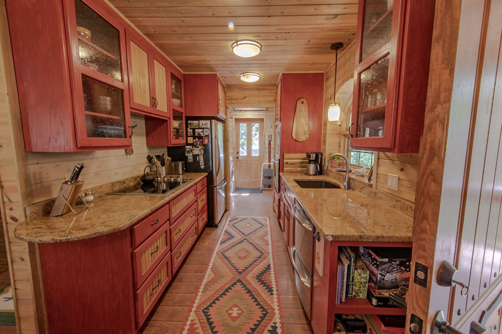 A narrow kitchen with wooden cabinets, granite countertops, and a patterned rug.