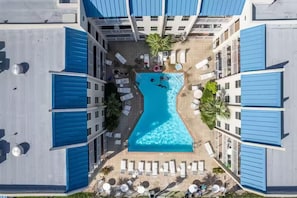 Eine Vogelperspektive auf ein Hotel mit einem zentral gelegenen Swimmingpool, umgeben von Liegestühlen und einer Palme.