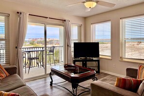 A living room with a sofa, a coffee table, a television, and a balcony with a view of the beach.