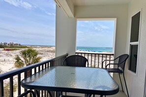 A balcony with a glass table and chairs, overlooking a beach with a clear sky.