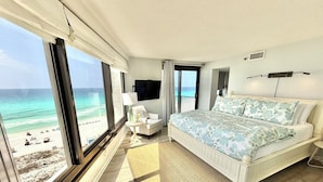 Camera da letto con una grande finestra, un letto, una sedia, una TV e vista sulla spiaggia.