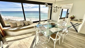 Zona pranzo moderna con tavolo in vetro, sedie bianche e vista sulla spiaggia e sull'oceano.