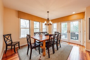 A dining room with a wooden table and chairs, large windows, and a chandelier.