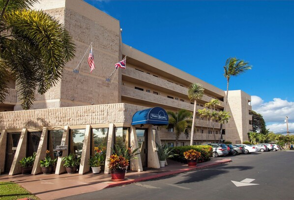 A hotel with a modern design, featuring a prominent entrance with a blue awning and the name 'Maui Sunset'. There are multiple flags, including the American flag, and several potted plants along the sidewalk.