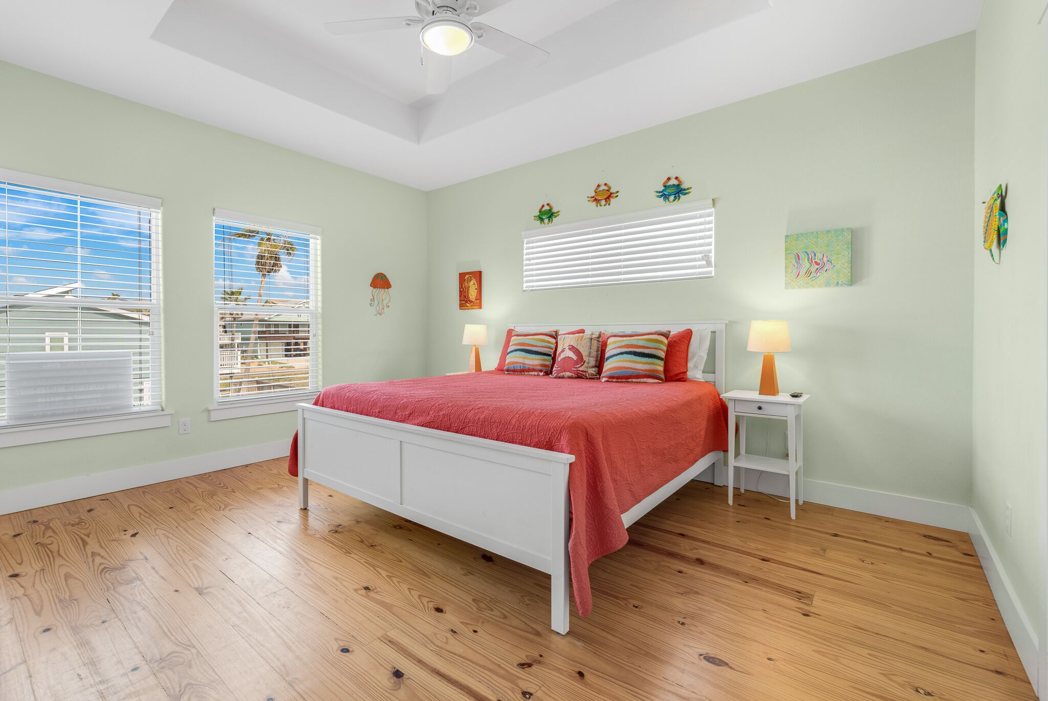 A bedroom with a white bed, two bedside tables with lamps, a ceiling fan, and a view of the outdoors through the windows.