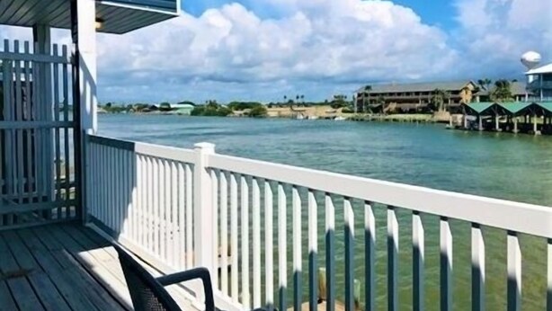 A waterfront view from a balcony with a white railing, overlooking a calm body of water and a distant shoreline with buildings. - Three Bedroom, Three and a Half Bath. Private Boat Slip. Communal Pool. (Port Mansfield)