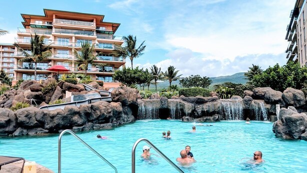A swimming pool with a waterfall feature, surrounded by rocks and palm trees, with a hotel building in the background.