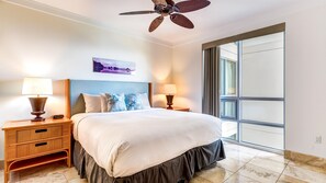 Une chambre d’hôtel avec un lit, deux lampes de chevet, un ventilateur de plafond, une grande fenêtre et une œuvre d’art accrochée au mur.