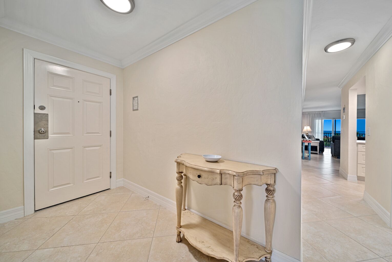 A hallway with a white door, a small table with a bowl, and a view of a balcony with a table and chairs.