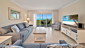 Ein modernes Wohnzimmer mit grauem Sofa, einem Couchtisch, einem gestreiften Sessel, einem Fernseher auf einem Holzboden und einem Balkon mit Blick auf den Strand.