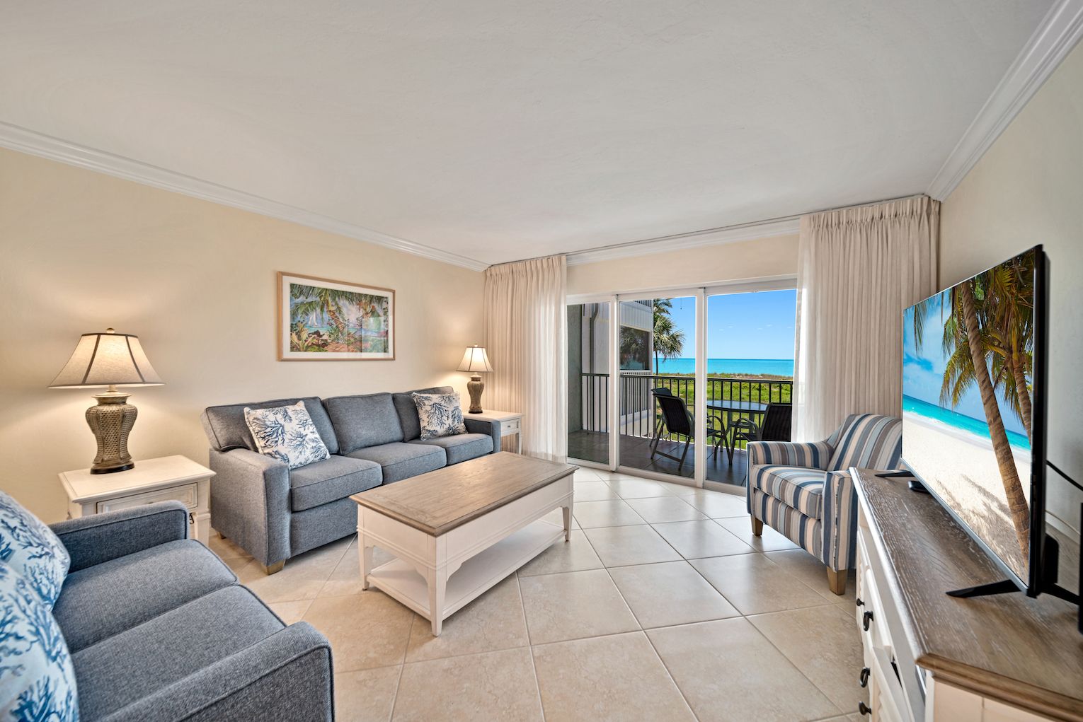 Un vaste salon comprenant un canapé, une table basse et un téléviseur posé sur un meuble en bois. Un balcon avec vue sur la plage est visible à travers les portes vitrées.