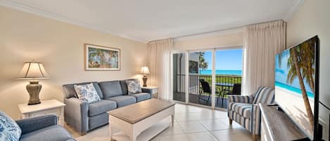 A spacious living room with a sofa, coffee table, and a TV on a wooden cabinet. A balcony with a view of the beach is visible through the glass doors.