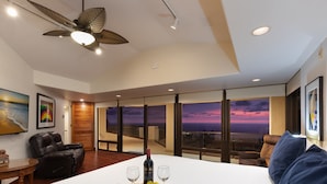 Habitación de hotel con una cama grande, ventilador de techo, vista al atardecer y una mesa de comedor con una botella de vino y copas.