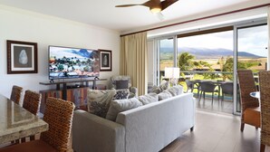 A spacious living room with a large flat-screen TV, comfortable seating, and a balcony with a scenic view.
