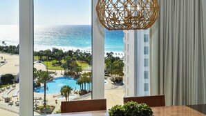 Habitación de hotel con un ventanal grande que da a una piscina y una playa, una lámpara colgante de tejido y vistas de un edificio al otro lado del agua.