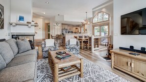 Een ruime woonkamer met een open haard, een eethoek met een houten tafel en stoelen, een keuken met moderne apparatuur en een groot raam met uitzicht op de omgeving.