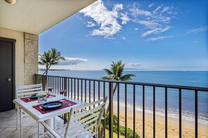 A balcony with a table set for two, overlooking a beach with palm trees and the ocean.