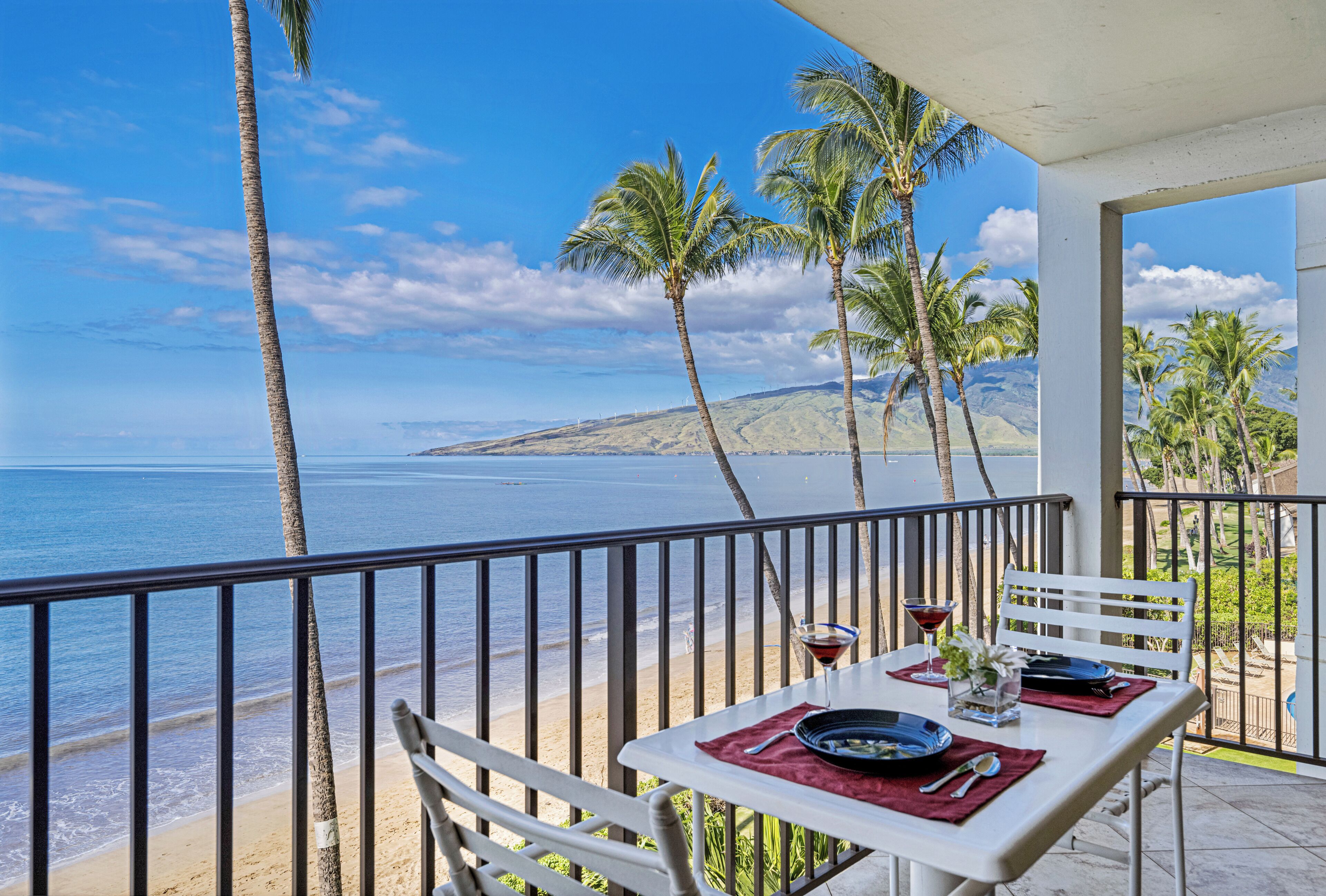 Un balcon avec une table dressée pour deux personnes, donnant sur une plage bordée de palmiers et offrant une vue sur l’océan.