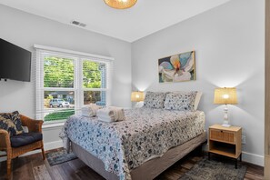 Une chambre à coucher avec un lit, deux lampes de chevet, une télévision, une fenêtre avec des stores et un tableau.