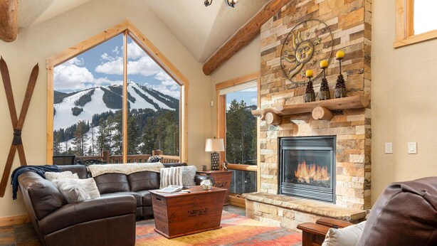 Un salon chaleureux avec une cheminée, des canapés en cuir, une table basse et une grande fenêtre donnant sur une montagne enneigée.