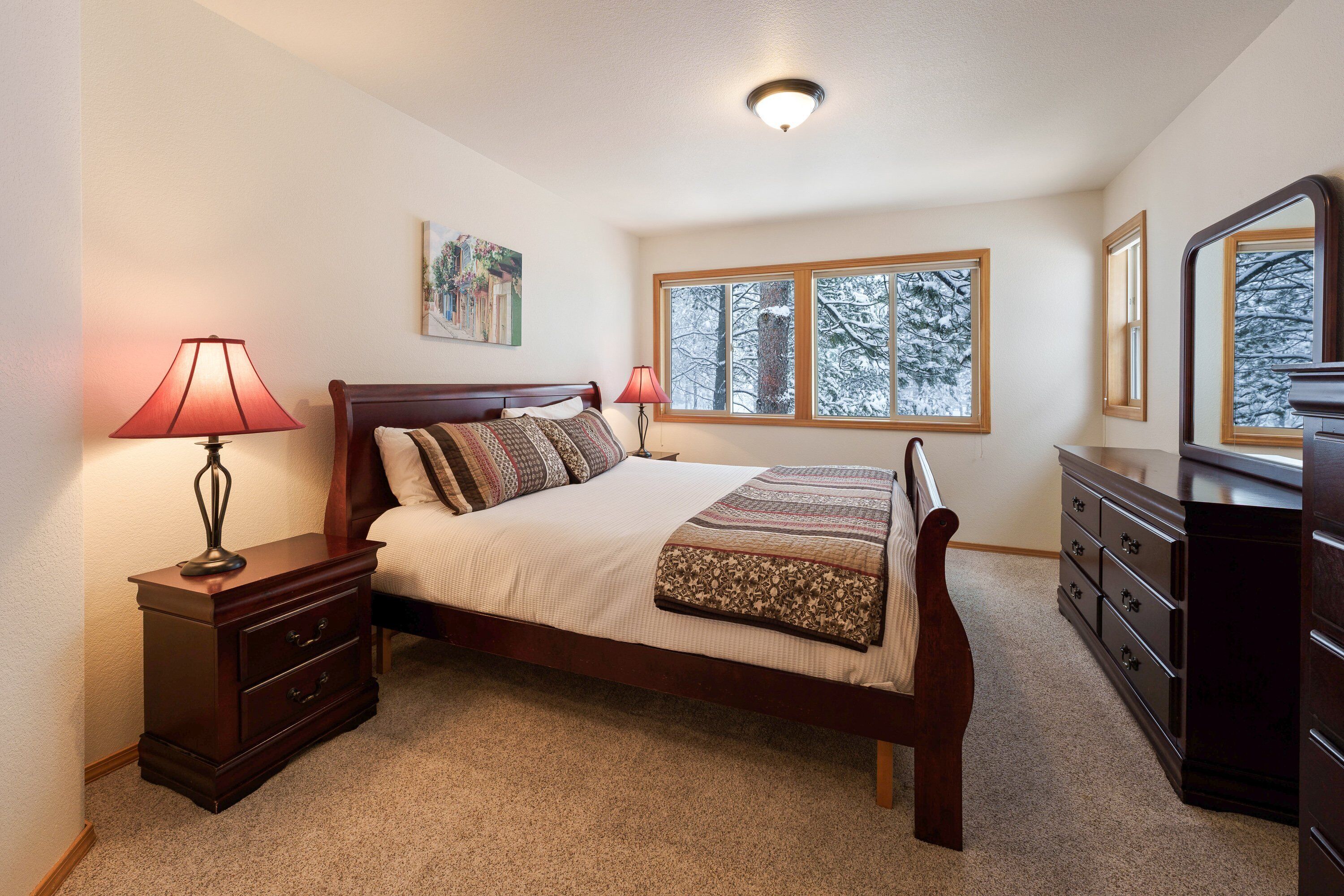 A bedroom with a bed, nightstand, dresser, and a window with a view of snow-covered trees.
