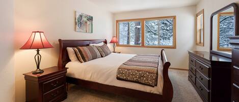 A bedroom with a bed, nightstand, dresser, and a window with a view of snow-covered trees.