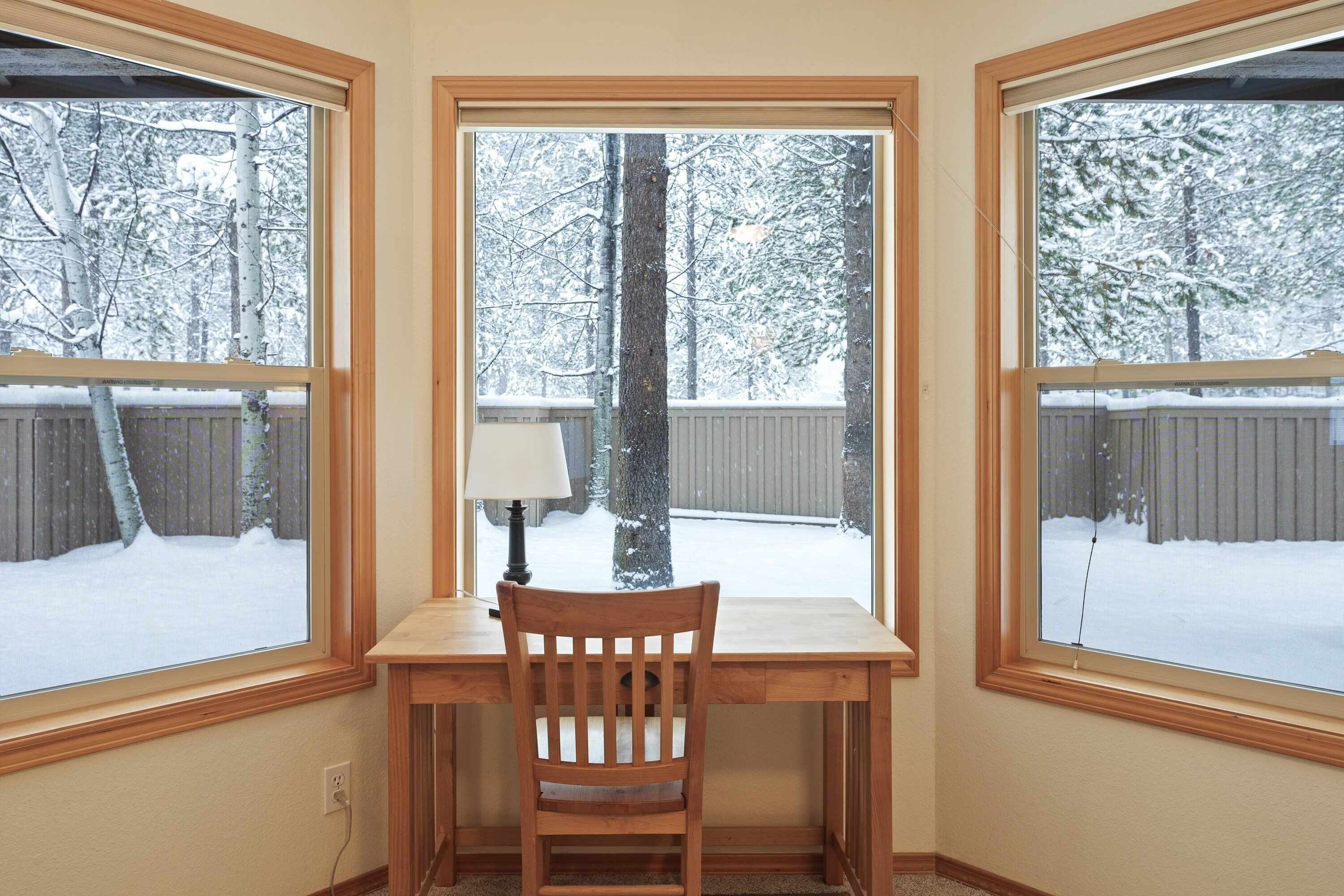 A corner room with a wooden desk and chair, a lamp, and a window overlooking a snow-covered backyard.