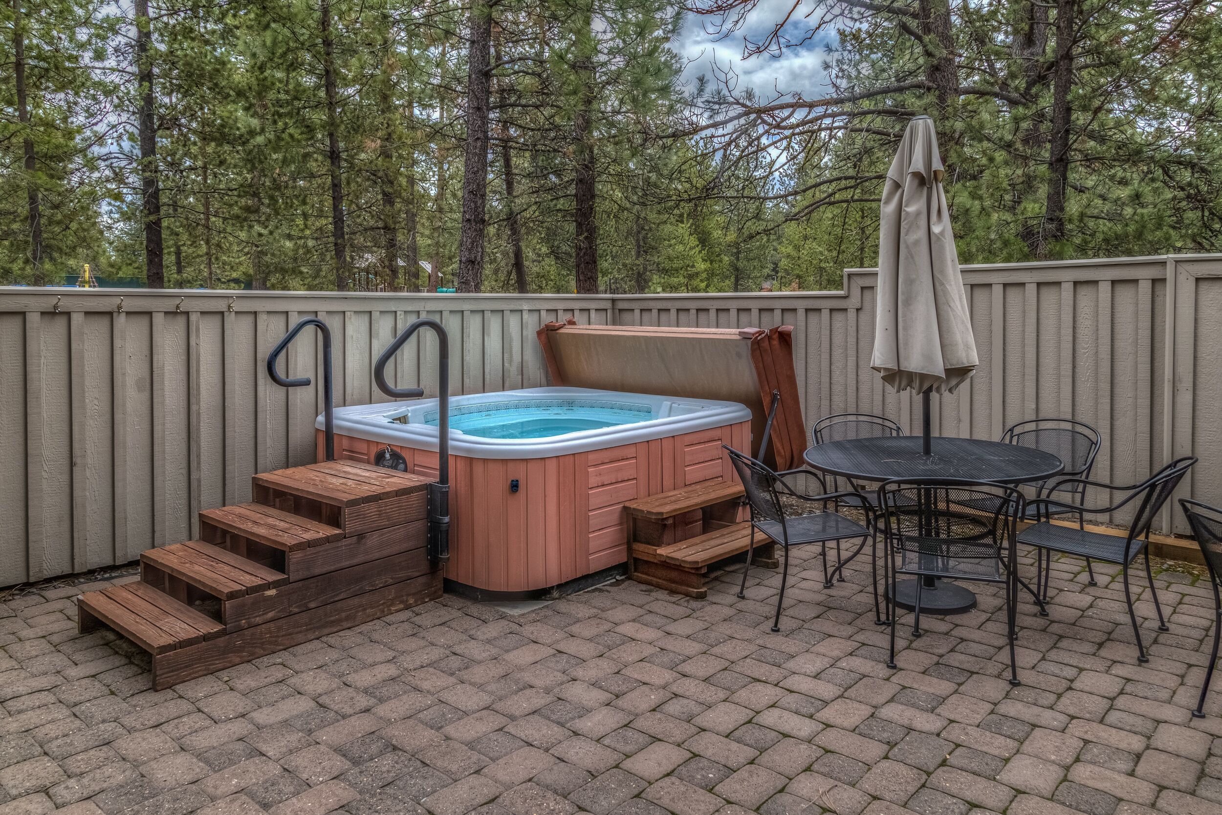 Un bain à remous avec des marches en bois et une table entourée de chaises, situés dans un jardin avec une clôture en bois et des arbres en arrière-plan.