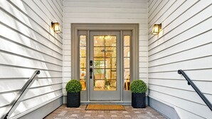 Une entrée avec une porte grise, deux jardinières noires ornées de plantes vertes et une allée pavée de briques.