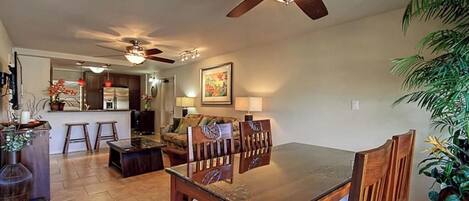 A dining area with a wooden table and chairs, a ceiling fan, and a kitchen in the background.
