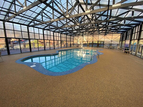 An indoor swimming pool with a glass ceiling, surrounded by a pebble floor and equipped with poolside chairs and tables.