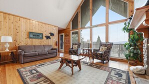 A cozy living room with a wooden floor, a brown sofa, patterned rugs, and large windows offering a view of the outdoors.