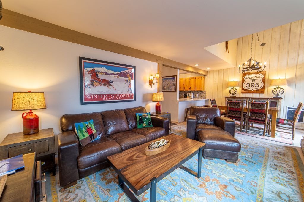 Una sala de estar acogedora con un sofá de cuero marrón, una mesa de centro de madera y un tapiz de colores. En la pared, hay un cuadro de una escena invernal y un letrero que dice "California US 395".