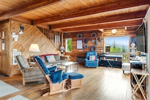 Interior acolhedor de uma cabana com decoração em madeira, um sofá, uma cadeira de balanço, uma escrivaninha e vista através de uma grande janela.