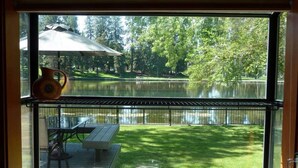 A view from a room with a large window overlooking a lake, a picnic table, and a glass table. - Mirror Pond - Cozy Cabin Directly on Mirror Pond - Deschutes River! (Bend)