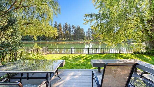 A deck with glass-top tables and chairs overlooking a lake, surrounded by trees. - Mirror Pond - Cozy Cabin Directly on Mirror Pond - Deschutes River! (Bend)