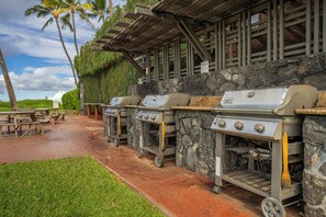 A row of barbecue grills with a stone wall backdrop and a covered seating area with picnic tables.