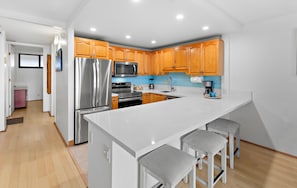 A modern kitchen with wooden cabinets, a stainless steel refrigerator, a microwave, and a large island with bar stools. - Beachy condo with garden and ocean views (Lahaina)