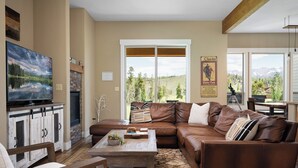 A cozy living room with a brown leather sofa, a wooden coffee table, and a television on a white cabinet. Large windows offer a view of a mountainous landscape.