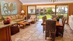 A spacious living area with a sofa, coffee table, and dining table set. There is a large window offering a view of the outdoors. - Ma'alaea Surf, Unit G10, 2BR Beachfront Condo, AC, Wi-Fi, Pool, TV, Kitchen (Kihei)